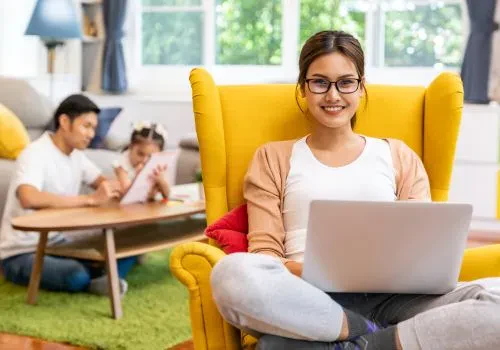 A stay-at-home mom smiling on her laptop in focus while a child and her father is in the blurred background